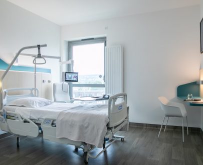 Chambre Institut de Réadaptation d'Achères avec lit simple médicalisé, fauteuil près d'une grande fenêtre, bureau et télévision.